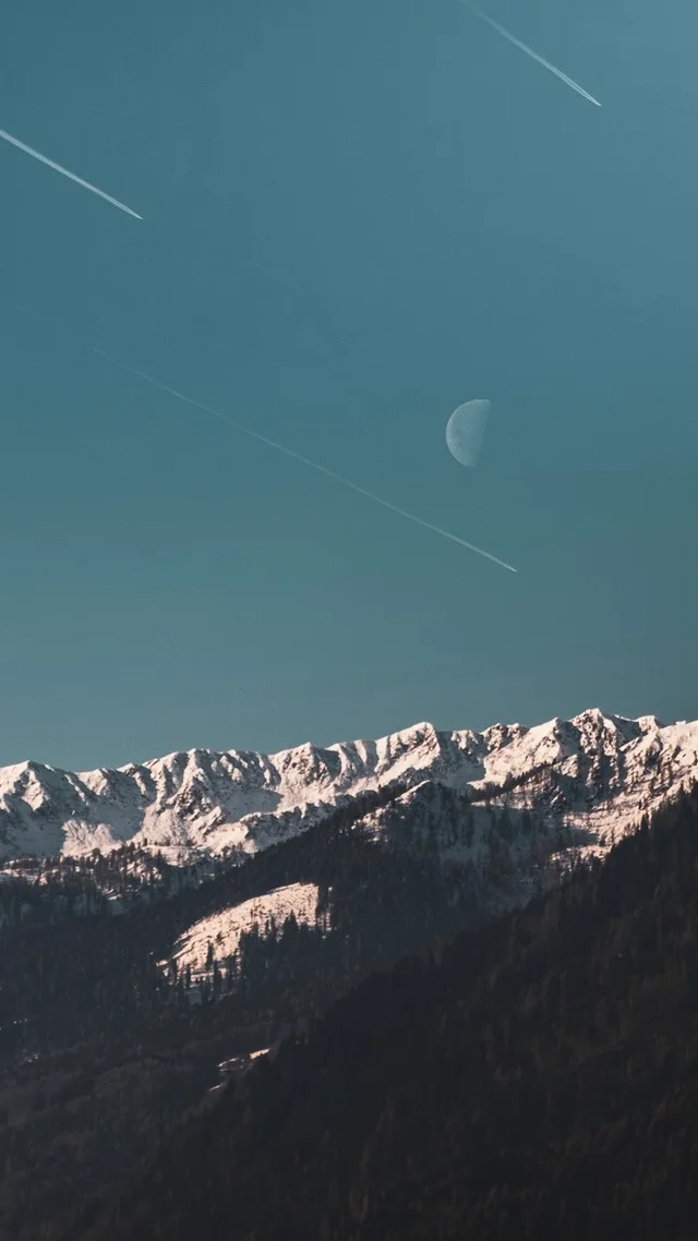 Snow-covered mountain peaks beneath the moon in a blue sky
